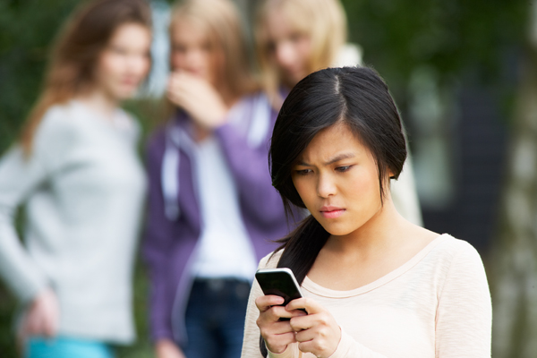 Teen girl with concerned look on her face looking at a mobile device, while in the background, a blurred group of teen girls is whispering and looking at the girl with the mobile device