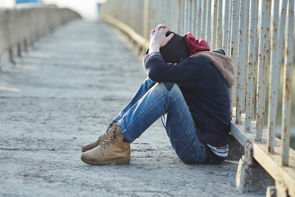 Young person alone, sitting next to a fence along a sidewalk, head down, hiding his or her face