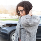 Woman standing in front of a two car crash, massaging the back of her neck, grimacing in pain.