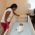 Woman looks over infant sleeping on its back in a crib that does not include soft or loose bedding, crib bumpers, toys, or other objects in the sleep area. 
