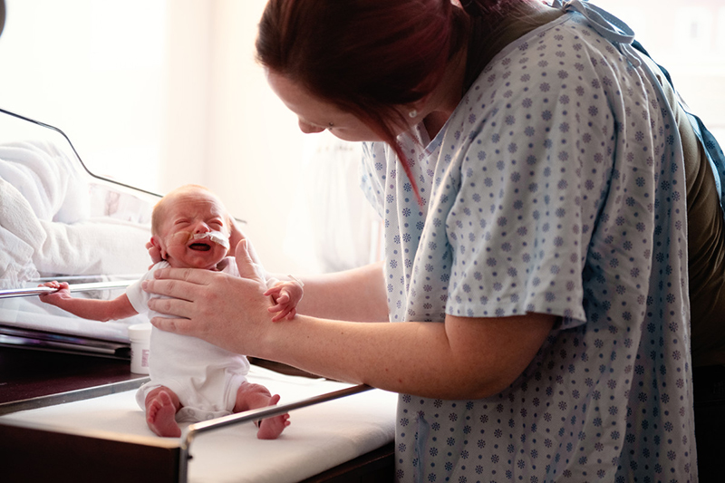 Woman in hospital gown supports preterm infant seated on table.