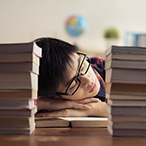 Teen male resting his head on a desk between two stacks of books.
