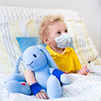 Masked child sitting in hospital bed holding a stuffed toy.
