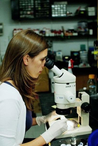 A woman with brown hair wearing a white jacket and white lab gloves peers into a microscope. Bottles and other scientific research equipment are blurred in the background.