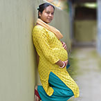 A pregnant woman wearing a yellow tunic, teal pants, and red sandals stands in an alley with her hands on her belly.