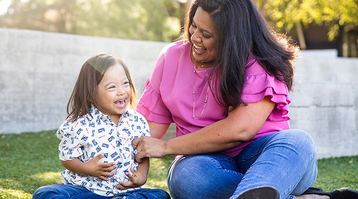 A little child with Down syndrome sits on the grass with their adult caretaker. Both are laughing. Trees and a stone wall are visible in the background.