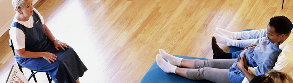 A pregnant woman and her partner sit together on a floor mat facing an instructor in a labor and delivery class. 