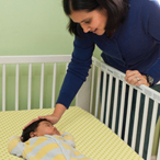 Mother next to crib, comforting sleeping baby with text: This is what a safe sleep environment looks like. The baby's sleep area has no bumpers, pillows, blankets, or toys and is next to where parents sleep.