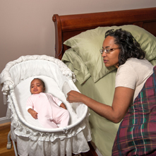 Room sharing: Mom in adult bed looks at on infant, who is sleeping in bassinet right next to the adult bed.