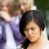 Teen girl with concerned look on her face looking at a mobile device, while in the background, a blurred group of teen girls is whispering and looking at the girl with the mobile device