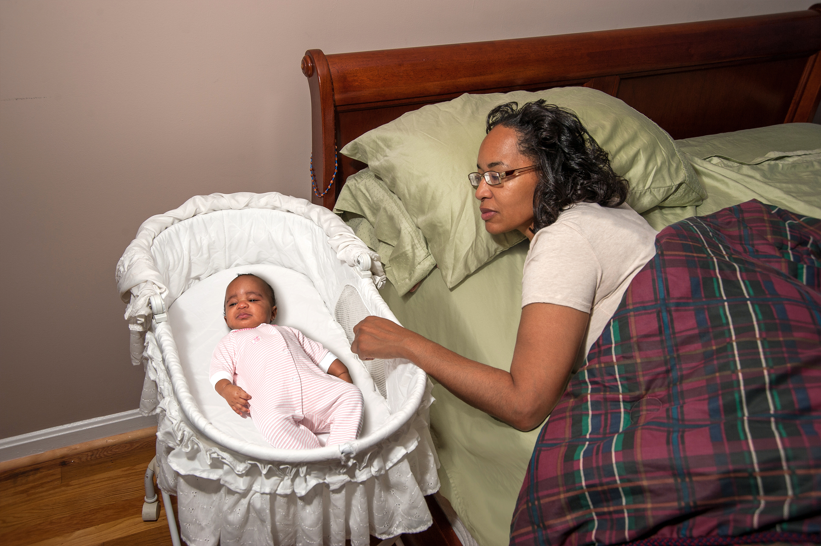 Room sharing: Mom in adult bed looks at on infant, who is sleeping in bassinet right next to the adult bed.