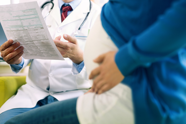 Midsection of pregnant woman pressing her hand on the side of her belly, with health care provider looking at folder in the background