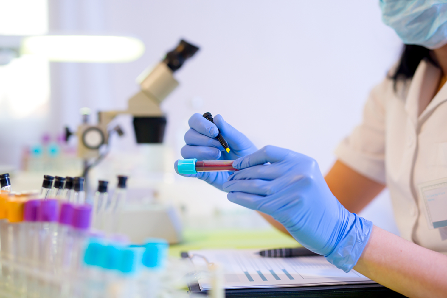 Researcher marking a test tube containing a blood sample in a medical laboratory, with a microscope in the background and other test tubes in the foreground.