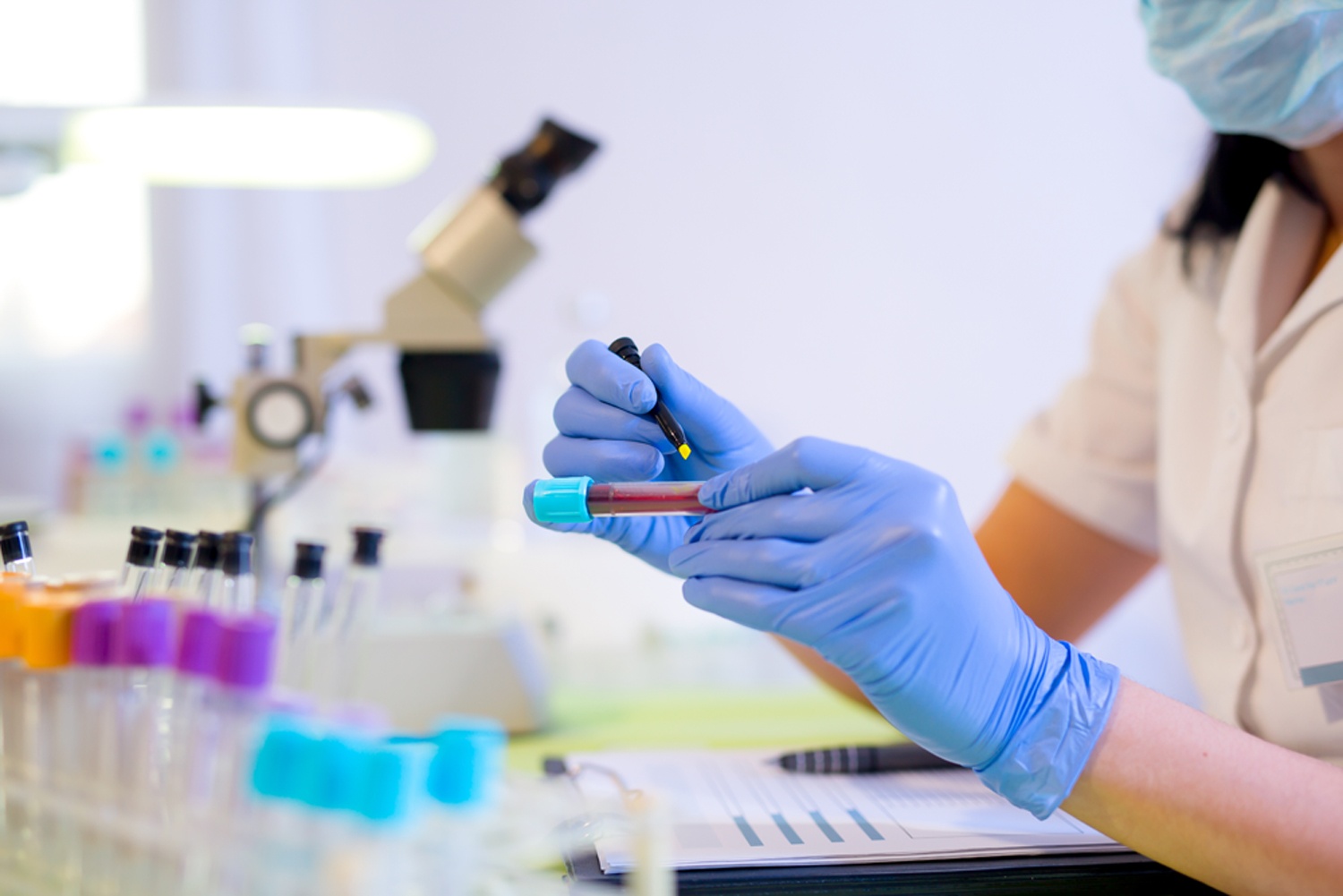 Researcher marking a test tube containing a blood sample in a medical laboratory, with a microscope in the background and other test tubes in the foreground.