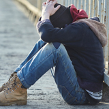 Young person alone, sitting next to a fence along a sidewalk, head down, hiding his or her face