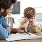 An adult male points to a notebook page and a child looks on, discouraged, with head in hands.