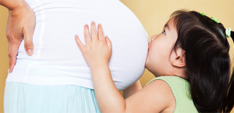 Stock image of a young girl kissing her pregnant mother’s stomach.
