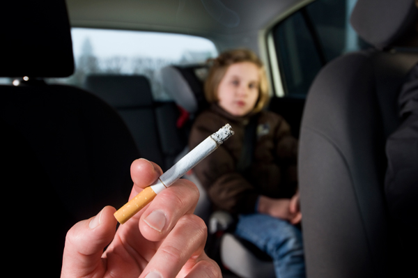 A hand holding a lit cigarette is visible inside a car with a young girl sitting in the backseat. 