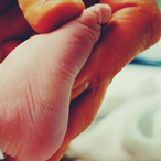 A hand grasps the foot of a newborn, which has an unmarked hospital ID tag around it.