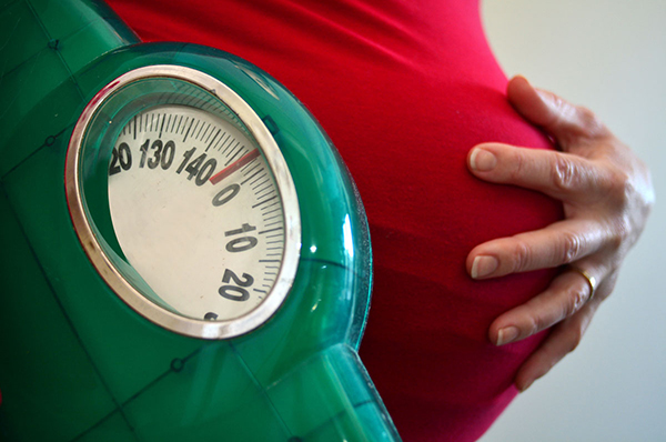 Pregnant woman holding a bathroom-style scale in one hand while cupping her other hand over her stomach.