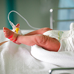Infant in a hospital crib, feet and torso visible, with medical devices attached to feet.