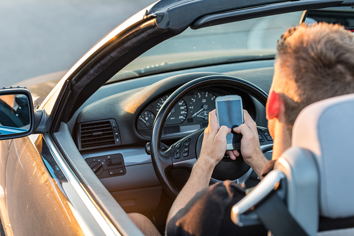 Stock image of a teenage boy using cell phone with two hands behind the wheel.