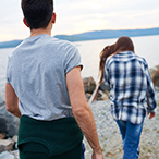 A young woman walking away from a young man along the shore of a river or lake.