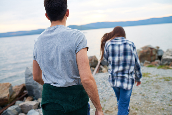 A young woman walking away from a young man along the shore of a river or lake.