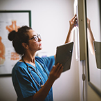 A doctor dressed in scrubs is examining an x-ray against a light box while holding a digital tablet.