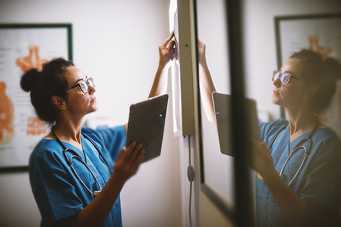 A doctor dressed in scrubs is examining an x-ray against a light box while holding a digital tablet.