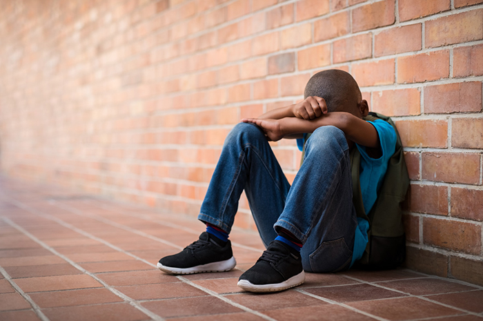 Child seated, leaning against brick wall, face resting on crossed arms.