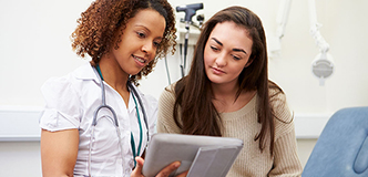 A young woman in a healthcare setting is looking at a digital tablet with her healthcare provider.