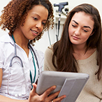 A young woman in a healthcare setting is looking at a digital tablet with her healthcare provider.