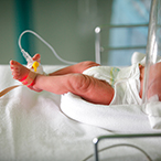 An infant in an incubator, with an oxygen sensor on one foot and an intravenous line on the other foot.