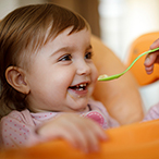 Baby girl in high chair smiling and being fed baby food on a spoon.