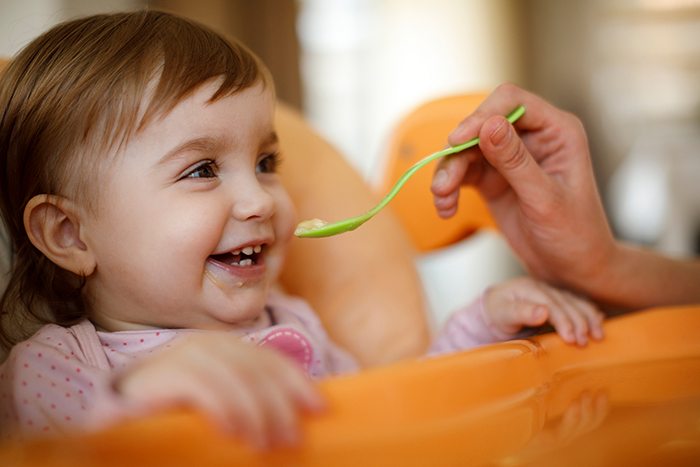 Baby girl in high chair smiling and being fed baby food on a spoon.