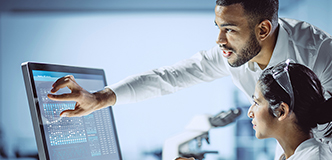 Two researchers in white lab coats, one seated and one standing, are looking at genetic data on a computer monitor.