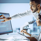 Two researchers in white lab coats, one seated and one standing, are looking at genetic data on a computer monitor.