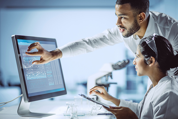 Two researchers in white lab coats, one seated and one standing, are looking at genetic data on a computer monitor.