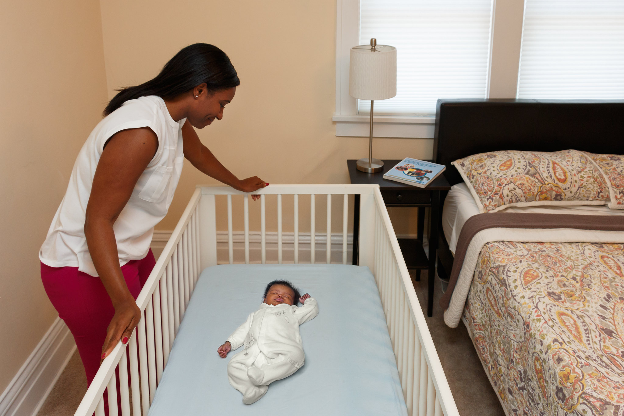 Woman looks over infant sleeping on its back in a crib that does not include soft or loose bedding, crib bumpers, toys, or other objects in the sleep area. 