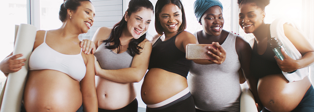 Cinco mujeres embarazadas sonrientes de distintos tipos de cuerpos, cuatro con sus abdómenes expuestos, se toman una "selfie". Embarazo para todas las tallas.