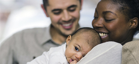 A man and woman sit together on a couch as the woman cradles a baby in her arms. Both are smiling at the baby.