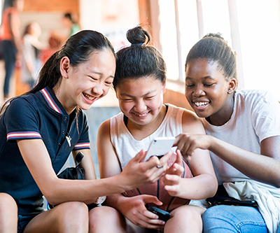Three teenaged girls laugh as they all look at one of their mobile devices.