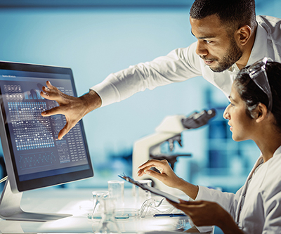 Two researchers in white lab coats, one seated and one standing, are looking at genetic data on a computer monitor.