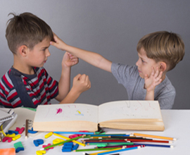 Two boys seated at table, one with fists clenched the other with hand on first child’s forehead.