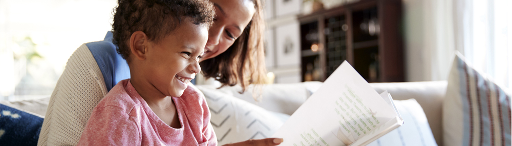 A little boy sits in a woman’s lap, laughing as she reads to him.