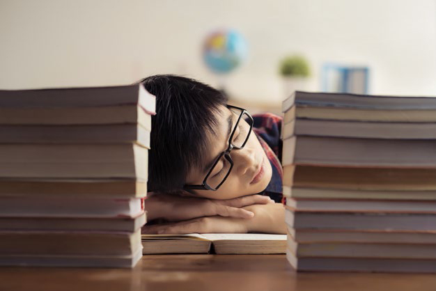 Teen male resting his head on a desk between two stacks of books.