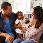 A smiling infant is held by her smiling parents, who are seated on a couch in their home.