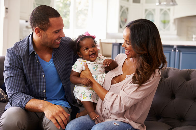 A smiling infant is held by her smiling parents, who are seated on a couch in their home.