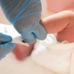 Gloved hands holding infant heel, blood spot visible on filter paper.